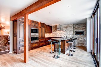 A modern kitchen with dark wood cabinets, a stone accent wall with a fireplace, a kitchen island with bar stools, and sliding glass doors.