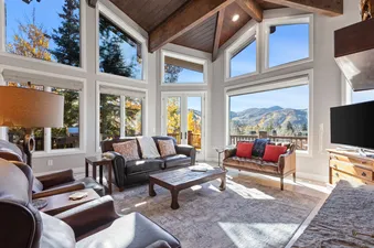 The living area at Bear Hollow 5428 features an impressive vaulted ceiling with exposed wooden beams, large windows offering panoramic views of the surrounding mountains and autumn foliage, and a comfortable seating arrangement with leather sofas and a ru