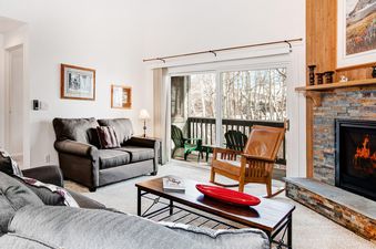 A cozy lounge area at Snow Flower 152 Park City featuring a fireplace with a stone hearth, a plush gray sofa, a rustic wooden coffee table, and a large glass door leading to a balcony with snowy trees outside.