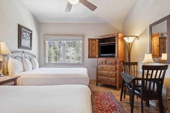 A cozy bedroom with two queen-size beds, a TV in a wooden cabinet, a dining table, and a window with a wooded view at The Charter B4305.