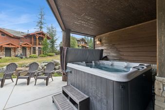 A large, private hot tub is shown on an outdoor patio, partially covered by a wooden overhang, with three Adirondack chairs beside it and cabin-style buildings in the background.