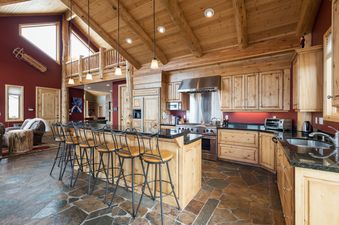 The kitchen at Solamere Home features a large wooden island with bar stools, natural wood cabinetry, stainless steel appliances, and a rustic stone floor.