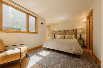 A spacious and inviting bedroom at Telluride Lodge Retreat features a large bed with a light-colored duvet and pillows, flanked by two nightstands with lamps, a window to the left with natural light, and a stylish leather armchair in the corner.