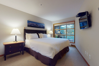 A cozy bedroom at Aspens on Blackcomb Unit 415 featuring a queen-sized bed, bedside table, lamp, and wall-mounted TV.