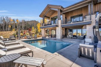 An expansive outdoor pool and patio area at The Sanctuary Chalet at The Colony, featuring chaise lounges and seating, with a grand chalet and autumn forest in the background.