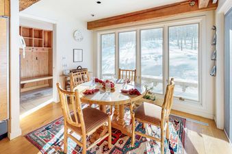 A round wooden dining table set for a meal, surrounded by wooden chairs, with large windows overlooking a snowy landscape in the dining area of the Trails Edge Vacation Home.