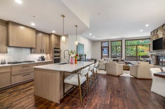 An expansive open-concept living space in Sunstone Chalet at One Beaver Creek features a modern kitchen with a large island and a comfortable lounge area with a stone fireplace and large windows showcasing outdoor views.