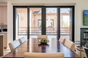 A dining room with a large wooden table, white chairs, and a view of a balcony with an umbrella through a sliding glass door.