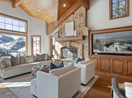 A spacious living room at Bellemont 5 featuring a stone fireplace, custom wooden cabinetry with a large television, plush seating, and stunning mountain views through large windows.