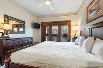 A spacious master bedroom in The Charter D340 with a king bed, dresser, wall-mounted TV, and a view of the outdoors through a set of double doors.