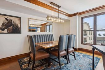 The dining area in Park Hyatt 4051 features a wooden dining table with a built-in banquette, complemented by upholstered chairs, all set on a patterned blue rug, with a large mirror and horse artwork adorning the wall.