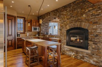 A rustic-inspired kitchen at The Palmyra Vista Cabin at See Forever, featuring a cozy stone fireplace, warm wood cabinetry, and a spacious island with granite countertops.