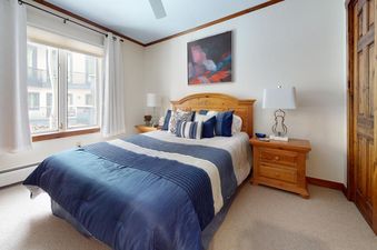 The All Seasons 3 Bedroom Platinum Unit B4 features a cozy bedroom with a large window offering a view of nearby buildings and a comfortable bed.