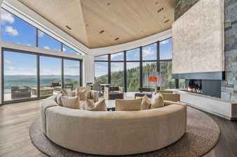 A luxurious and spacious lounge area inside Mountainhead, featuring a large circular sofa, a modern fireplace, and expansive windows with panoramic mountain and forest views.
