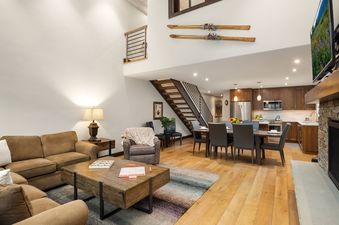 An expansive open-concept living area at Montaneros 402 features a comfortable lounge, a dining area, and a modern kitchen, all connected by natural wood flooring and a stylish staircase.