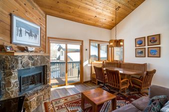 A cozy living and dining area at The Pines Condominiums A304, featuring a stone fireplace, wooden furniture, and glass doors opening to a snowy balcony.