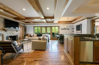 A luxurious open-concept living and dining area in Chateau Beaver Creek Residence 210, featuring a stone fireplace, comfortable sofas, a dining table, and large windows looking out onto green trees.