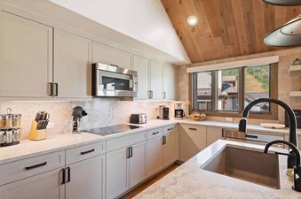 A modern kitchen with light gray cabinets, white marble countertops, and a wooden vaulted ceiling, featuring a sink, microwave, toaster, and coffee makers, with a large window showing a mountain view.
