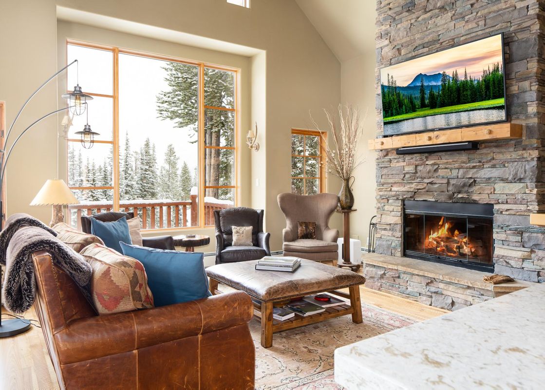 A cozy living room in Moonlight Mountain 38B Cowboy Heaven with a stone fireplace, comfortable leather seating, and large windows overlooking a snowy forest.