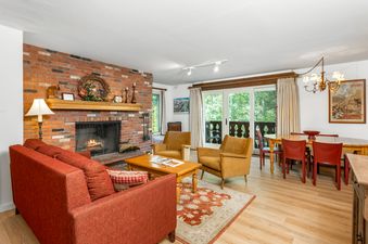 A cozy living room and dining area featuring a fireplace, comfortable seating, and a dining table, with large windows overlooking lush greenery.