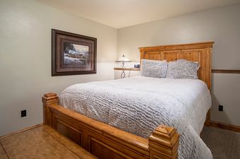 A cozy bedroom featuring a rustic wooden bed frame, a white textured comforter, and a framed winter-themed painting on the wall.