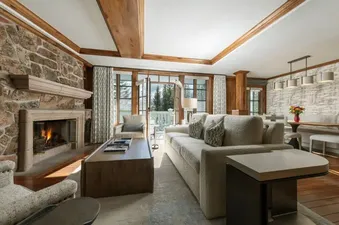 The spacious living room and dining area of Chateau Beaver Creek Residence 204 features a stone fireplace, comfortable sofas, and large windows with a view of snow-covered trees.