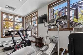 A bright and spacious gym area in Silver Strike Lodge 605, featuring various exercise equipment and large windows with wooden frames offering a view of the surrounding buildings and autumn trees.