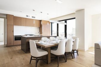 A modern kitchen and dining area with wooden cabinetry, white countertops, a large island, a wooden dining table with white chairs, and floor-to-ceiling windows.