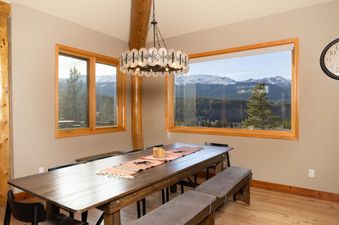 A rustic dining room features a long wooden table with benches, set against a backdrop of large windows offering stunning mountain views.