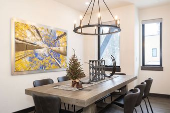 A rustic wooden dining table with a festive evergreen centerpiece and dark chairs beneath a modern chandelier in a well-lit dining room with large windows and a nature-themed painting on the wall.