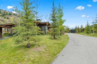 A modern cabin with dark wood siding and a flat roof is nestled among green grass, pine trees, and aspen trees under a blue sky, with a paved driveway leading to a detached garage.