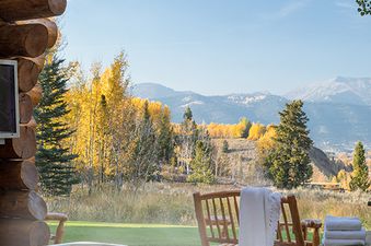 A private hot tub on a wooden deck offers stunning panoramic views of golden autumn trees and distant snow-capped mountains, complementing a rustic log cabin.