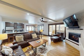 The living room at Phoenix 107 features a leather sofa, a coffee table with a cowhide rug, a fireplace, a large TV, and a view of the snowy outdoors through a window.