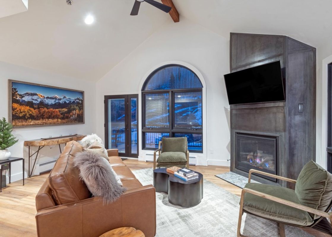 The living area of Le Chamonix Unit A features a large brown leather sofa, two green armchairs, a television mounted above a modern fireplace, and expansive windows offering a view of the snowy landscape.