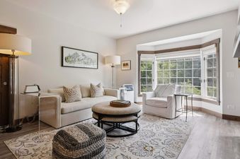 A bright living room at The Charter D3505 features a light-colored sofa with throw pillows, a round ottoman, and a large window with an outdoor view.