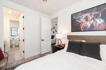 A modern bedroom at Alpenglow Retreat features a bed with white linens and dark pillows, a wooden headboard, a bedside table with a lamp, and a large, colorful painting of a highland cow, with a glimpse of the en-suite bathroom through an open door.