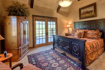 A luxurious bedroom at Antler Ridge Lodge featuring a large bed with ornate wooden frame, vibrant bedding, bedside lamps, a wooden armoire, and double glass doors leading to a balcony.