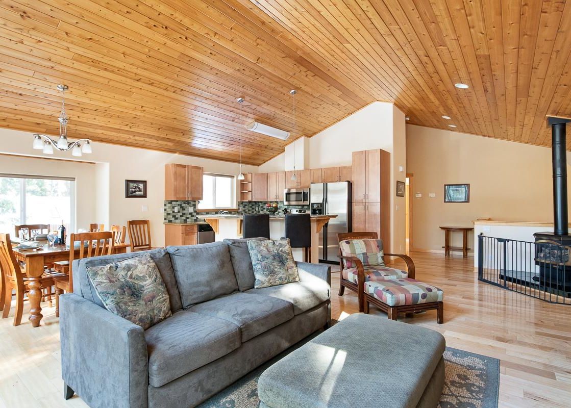 A wide view of the open-concept living space in North Shore Golden Pine, featuring a living room, dining area, and kitchen under a vaulted wooden ceiling, with a wood-burning stove to the right.