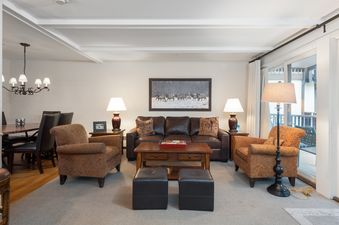The living area features a large brown leather sofa, two patterned armchairs, a wooden coffee table, and two black ottomans, with a dining area visible in the background and natural light from a large window.