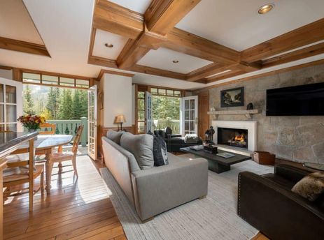 A spacious living and dining area in Chateau Beaver Creek Residence 305 with wood floors, a stone fireplace, comfortable seating, and French doors opening to a balcony with mountain and forest views.