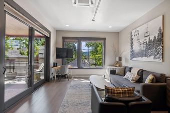 A modern living room with a grey sofa, coffee table, and desk, featuring large windows with views of trees and mountains.