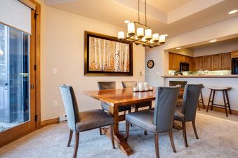 A sophisticated dining area featuring a wooden table with six upholstered chairs, illuminated by a rectangular chandelier, with a forest artwork on the wall and a partial view of the kitchen.