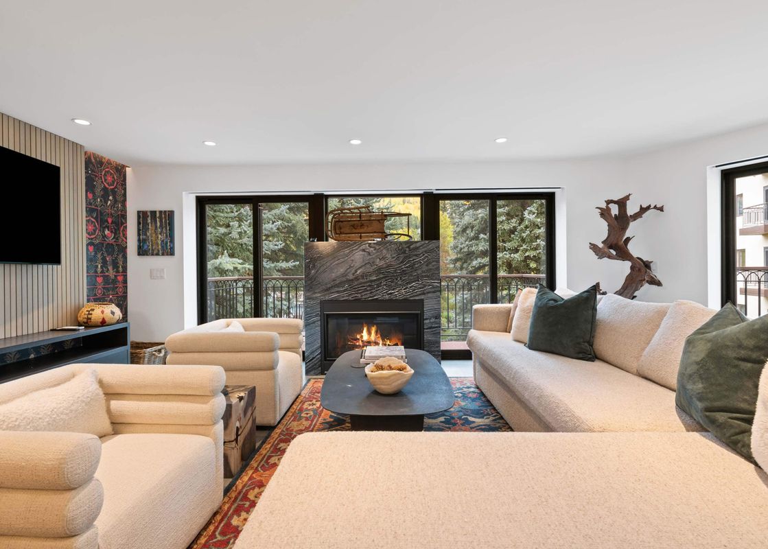 A modern and cozy living room with cream-colored sofas, a dark coffee table, and a fireplace, overlooking a scenic view of evergreen trees through large windows.
