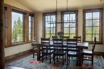A rustic dining room features a large wooden table with eight chairs, centered under a black metal chandelier, with multiple windows offering views of mountains and pine trees.