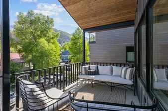 The balcony at Mears Station B features comfortable outdoor seating with a sofa, chair, and woven rug, all overlooking lush green trees, distant mountains, and charming buildings under a bright sky.
