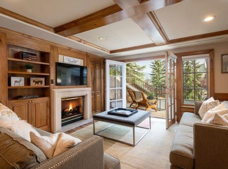 The Chateau Beaver Creek Residence 103's living room, featuring a cozy fireplace, a large television, comfortable sofas, and elegant wooden furnishings, with French doors opening to an inviting outdoor patio amidst lush greenery.