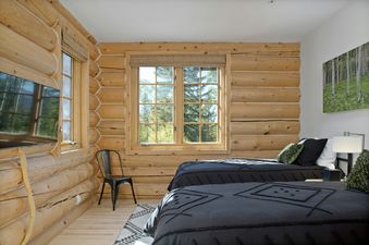 A cozy bedroom at Sundance Lodge 108 Lodges Lane features two beds with dark bedding, natural wood log walls, and a large window overlooking green trees.