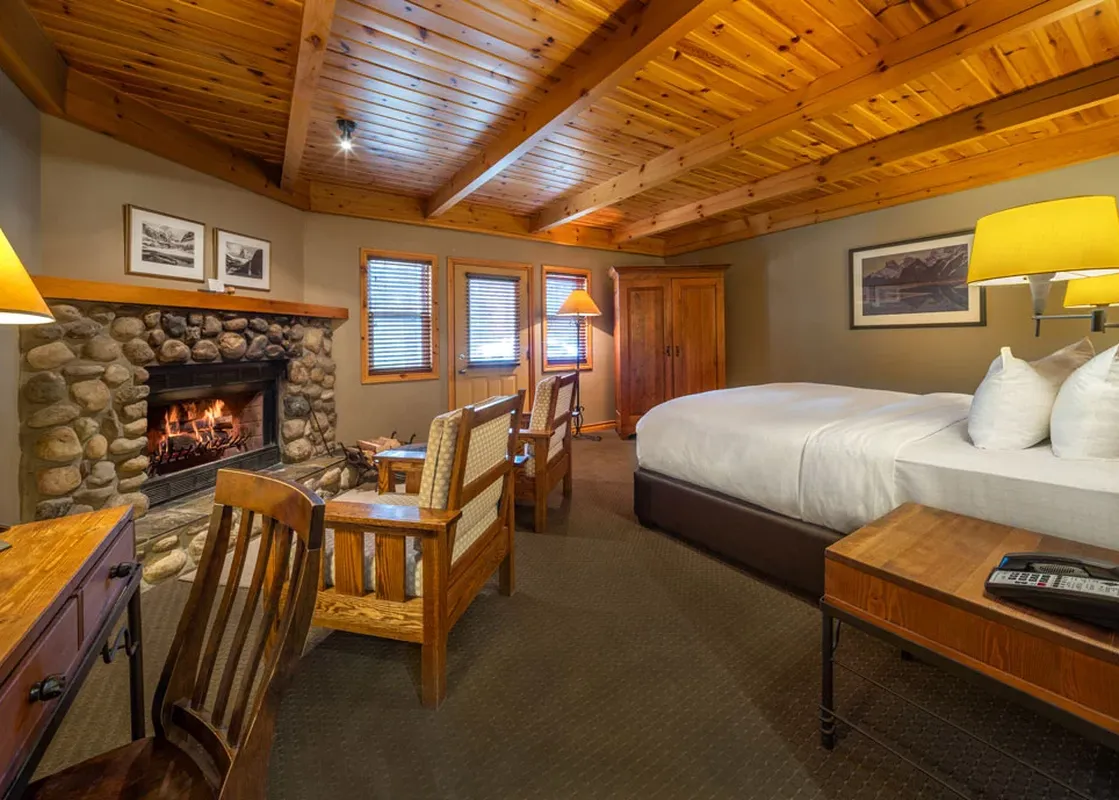 A cozy Buffalo Mountain Lodge Premier Room featuring a stone fireplace, a large bed with white linens, rustic wooden furniture, and wooden beamed ceilings.