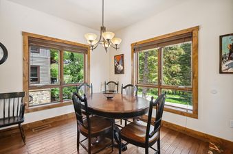 A dining area at Whitefish Ski Haus with a round wooden table and six chairs, featuring large windows with views of green trees and a neighboring house.