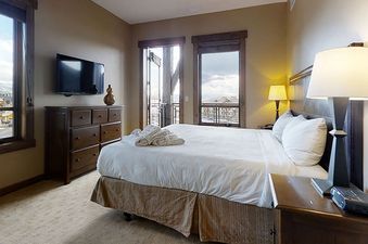 A cozy bedroom with a queen-sized bed, warm lighting, a flat-screen TV, and two windows offering snowy outdoor views.